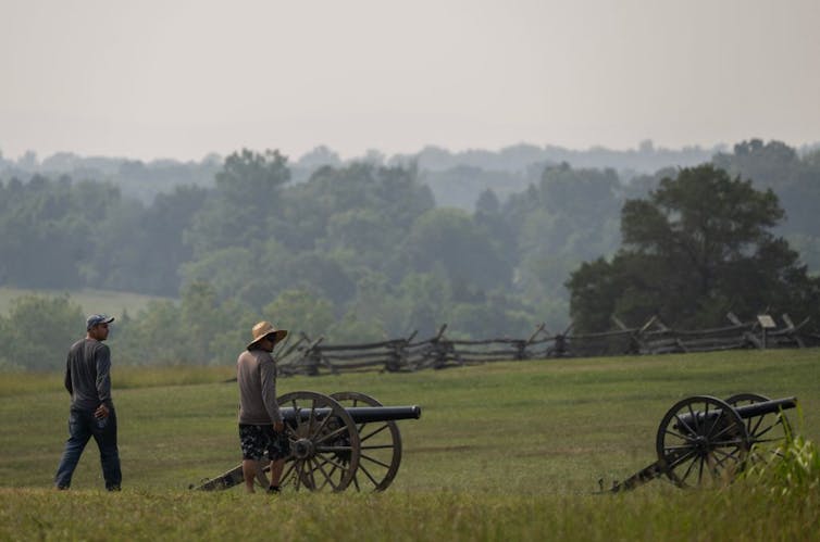 Two men walk across a green field with two portable cannons.