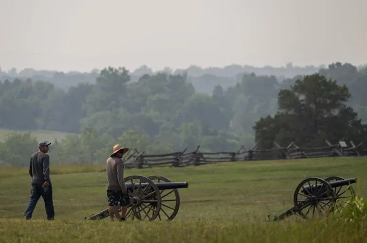 Two men walk across a green field with two portable cannons.