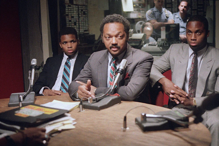 A Black man speaks in a radio studio as two younger Black men sit on either side of him.
