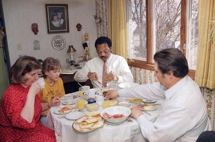 A Black man tucks into breakfast at a dining table with a white man, a white woman and their young boy.