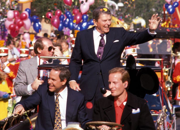 Colorful balloons surround a vintage car transporting four men wearing suits.