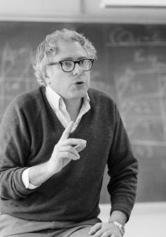 A man with wavy grey hair and glasses and wearing a wool sweater gestures as he speaks.