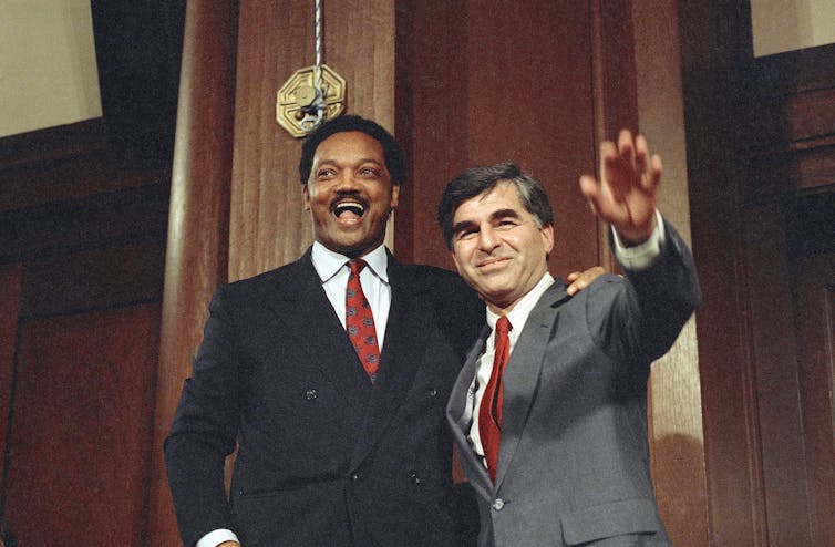 A dark-haired white man waves to photographers as a laughing Black man stands beside him.