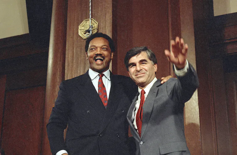 A dark-haired white man waves to photographers as a laughing Black man stands beside him.