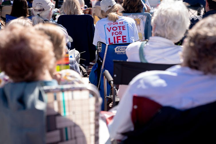 A crowd of people are seen sitting in folding chairs, including one person whose back of the shirt says 'Vote for life'