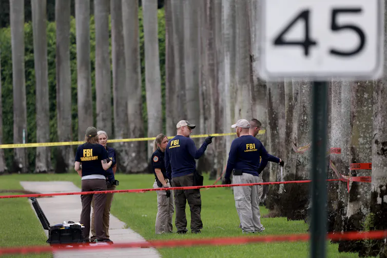 People wearing dark shirts stand behind orange police tape on green grass.