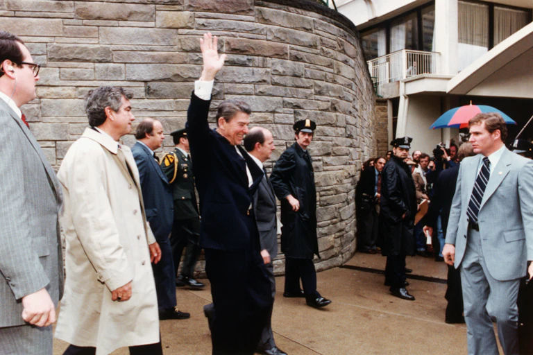 A man wears a black suit and waves his hand in the air, surrounded by other people wearing formal clothing in a vintage looking colored photo.