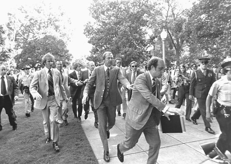 Men in suits and police uniforms are seen rushing together on a street in a black and white photo.