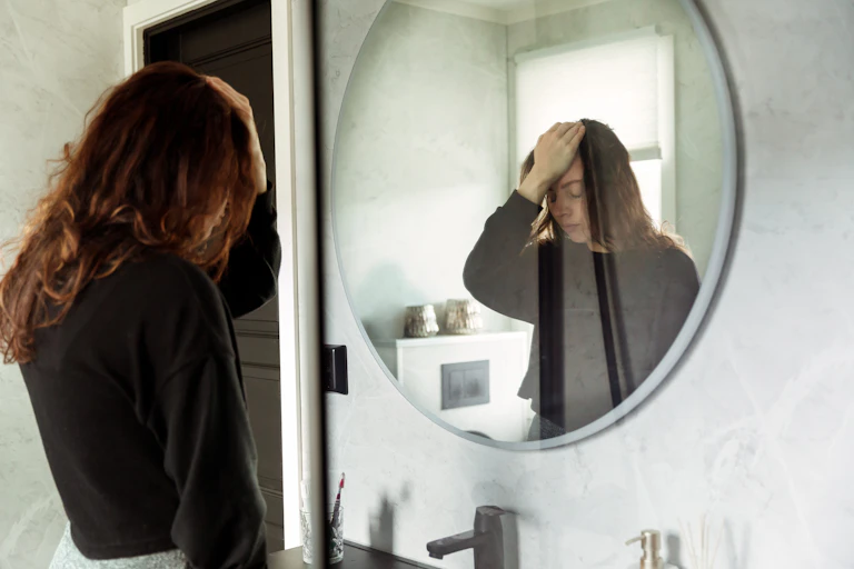 A young woman with long brunette hair clutches her forehead and looks downcast in front of a mirror.