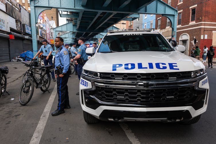 Uniformed police officers stand beside a patrol car under an elevated train