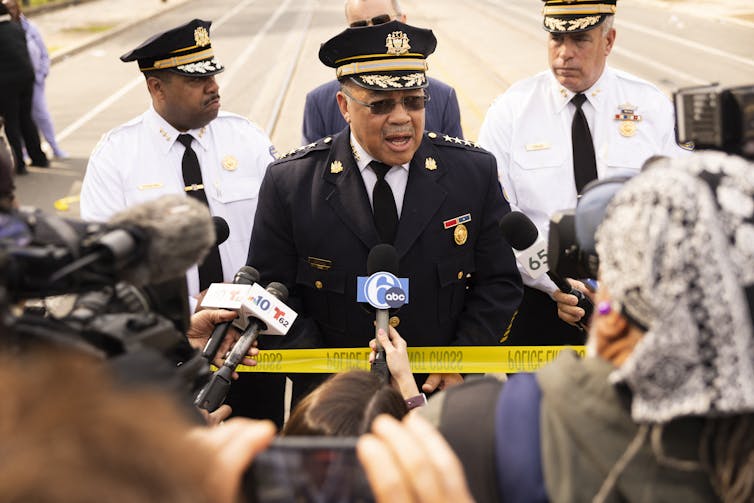 Police officer in uniform speaks at podium to people holding microphones