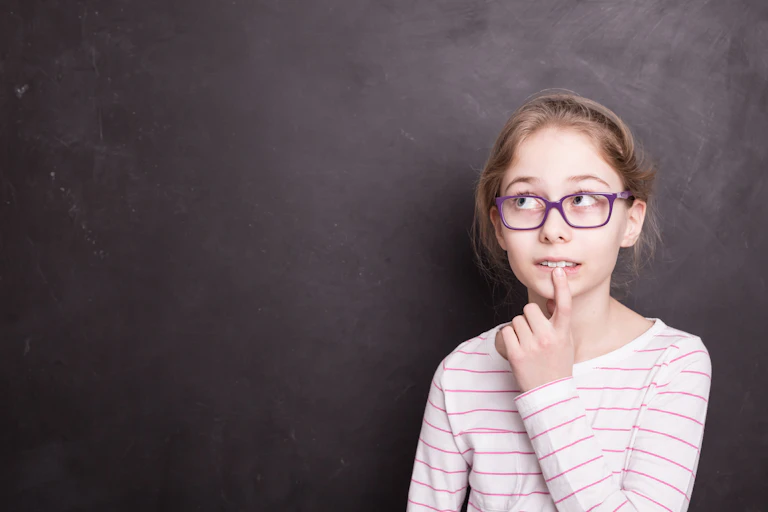 Uma menina de cerca de 10 anos em frente a um quadro negro de escola com expressão de dúvida