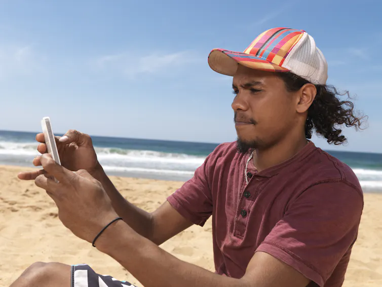 A young Aboriginal man on a beach looks at his phone.