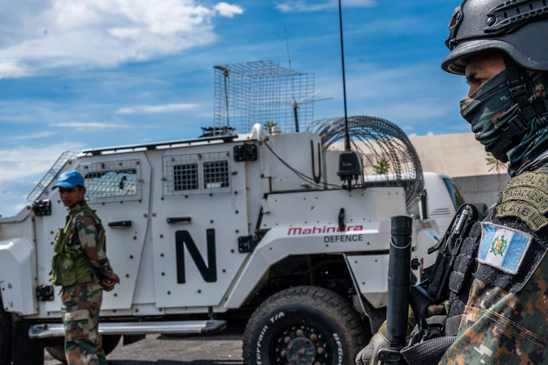Person in camouflage uniform and helmet, with armoured vehicle in the background
