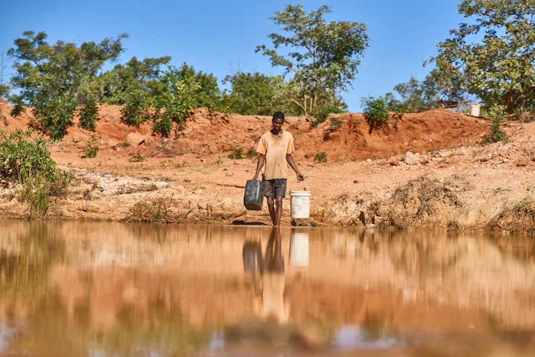 A man walks across dry earth in high temperatures to fetch water in buckets from a shallow and muddy pool