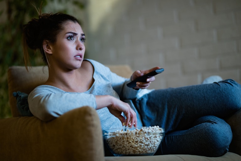 Woman watches TV and eats popcorn, looking sad