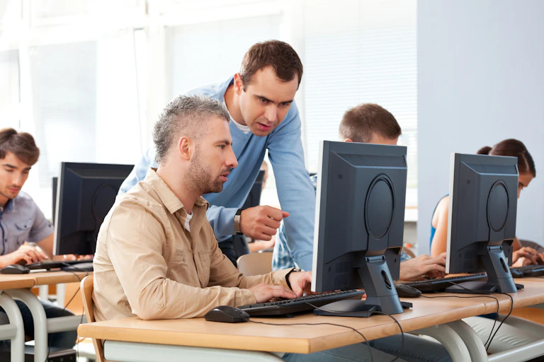 A man sits at a computer on a desk in a classroom while male teacher looks over his shoulder.