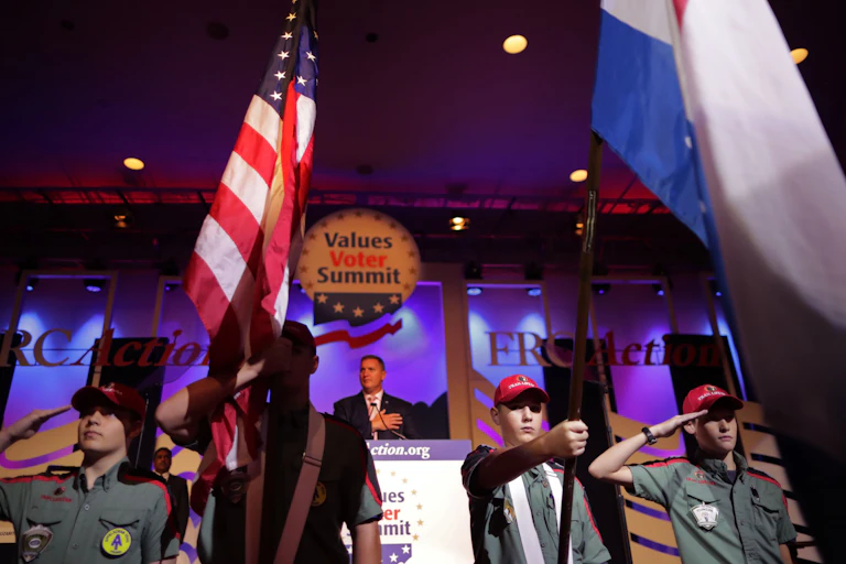 Boys hold flags and salute while wearing red caps below a sign saying 'Values Voter Summit.'
