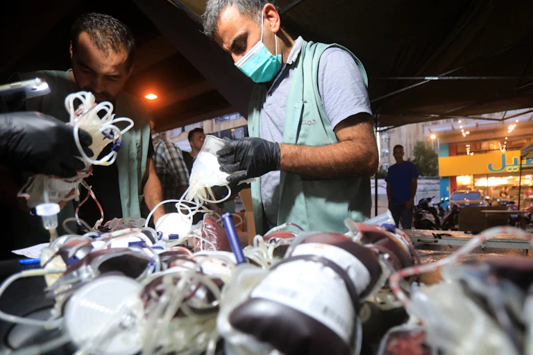 A man in a surgical mask and gloves holds tubes. In the foreground are bags of blood.
