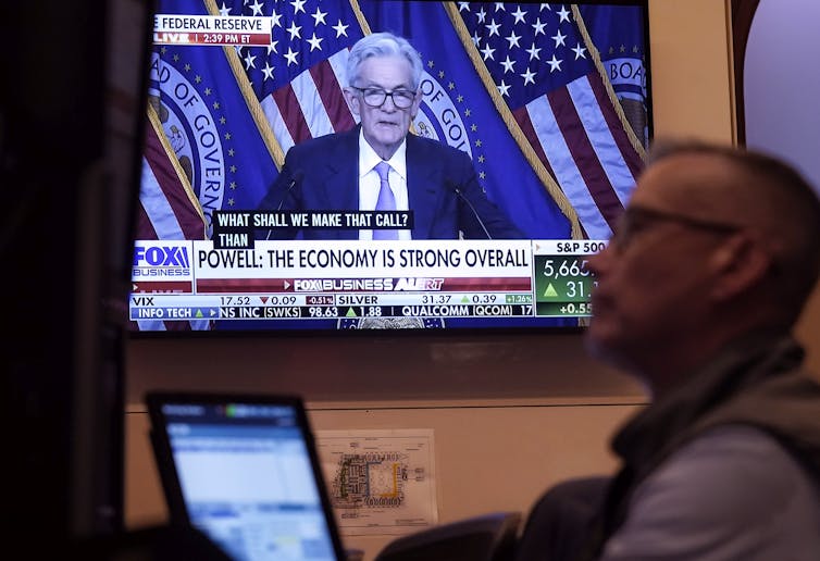 A trader sits at desk watching something as TV displays a fed news conference in background