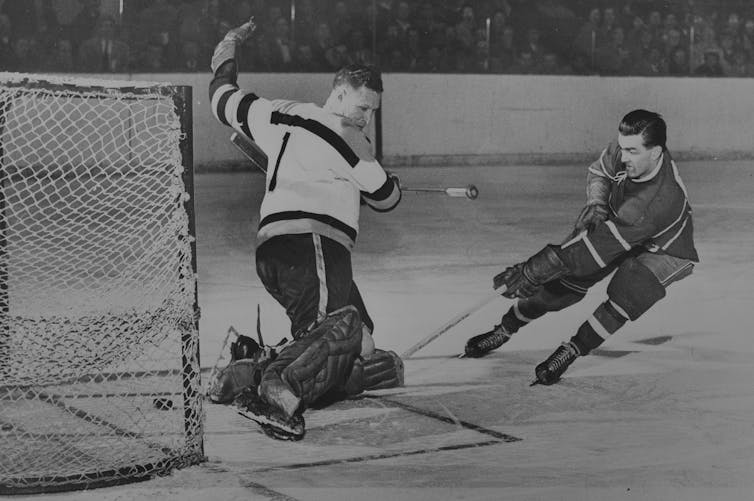 Black and white photo of a hockey player with no helmet scoring a goal against a goal who also doesn't have a helmet on