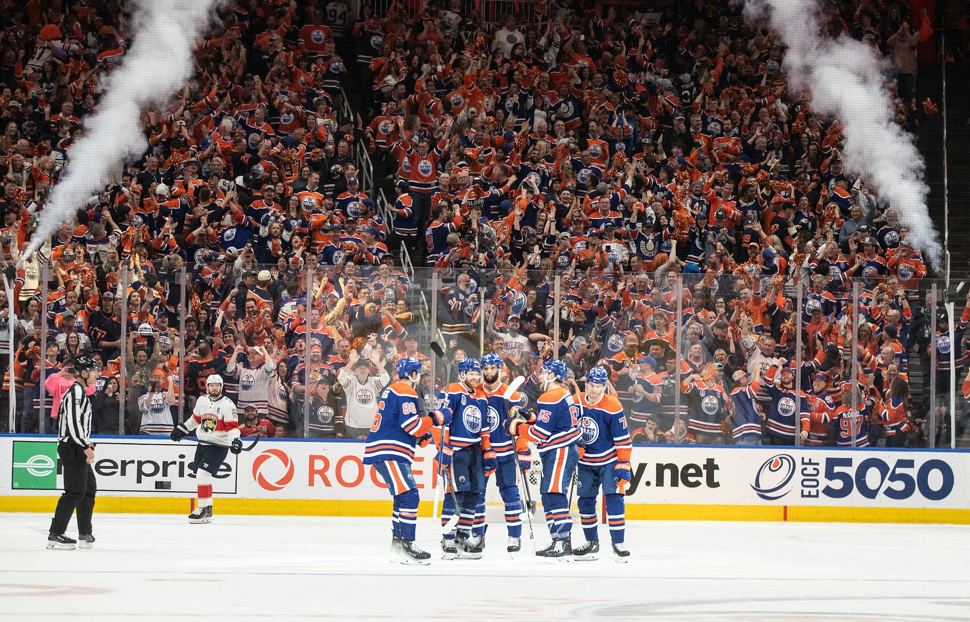 A group of Edmonton Oilers hockey players huddle in the middle of the ice. Behind them, the stands are packed full of people in blue and orange Oilers jerseys