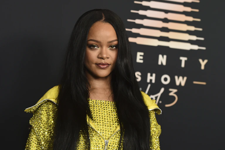 A young Black woman with long, black hair and a glittery yellow top poses in front of a backdrop that says 'Savage X Fenty Show Vol. 3'