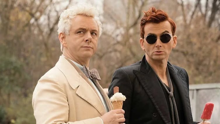 A middle-aged man in a beige suite and white, curly hair side eyes something off camera while holding an ice cream cone. Beside him stands a middle-aged man in a black suit with dark red hair and round sunglasses, holding a popsicle.