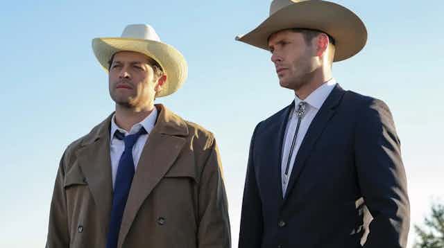 Two middle-aged men in suits and cowboy hats standing next to teach other. One is wearing a trench coat.