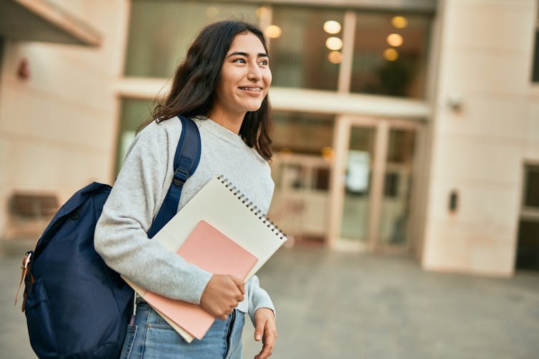 A young woman wears a backpack and holds notebooks outside a building.