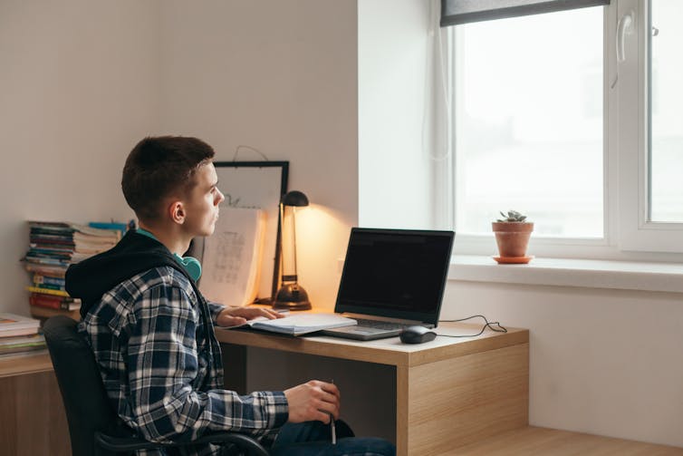 A young man sits at a desk, with a lamp and computer. He looks out the window.