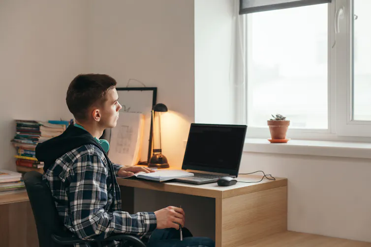 A young man sits at a desk, with a lamp and computer. He looks out the window.
