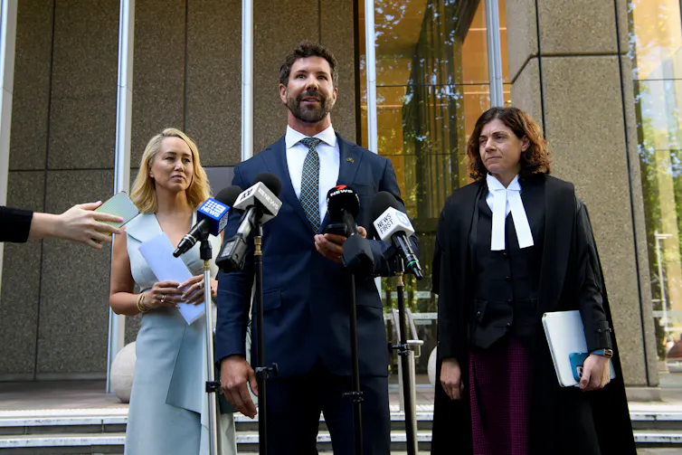A man in a suit stands in front of two women and addresses the media.