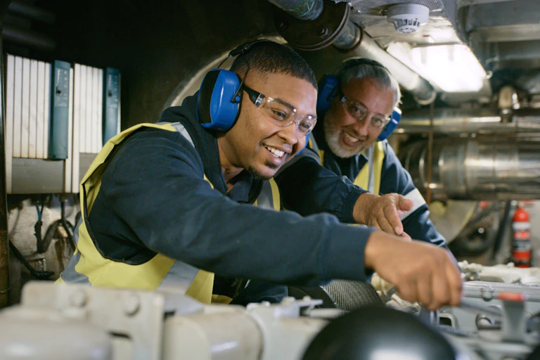 Smiling men in uniform working on engine repair in factory wearing safety glasses.