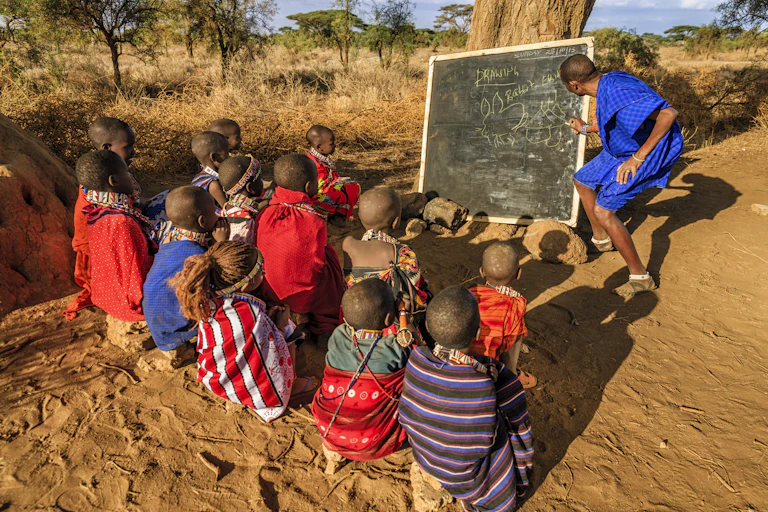 Children sit in front of a chalkboard under a tree outdoors