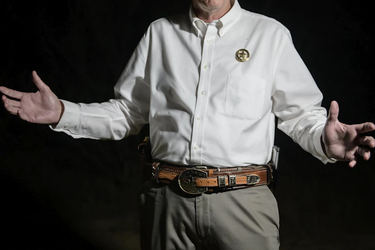A man wearing a badge and a leather belt gestures.