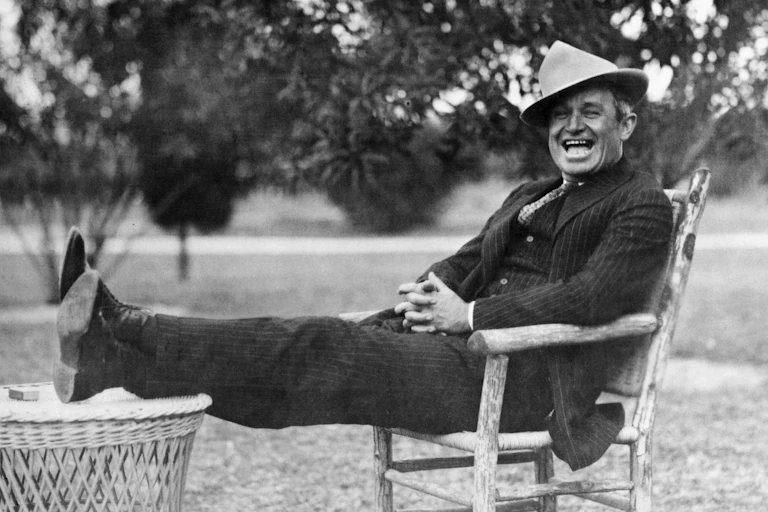 A man in a pin-striped suit with a funny hat on and his feet up, laughing.
