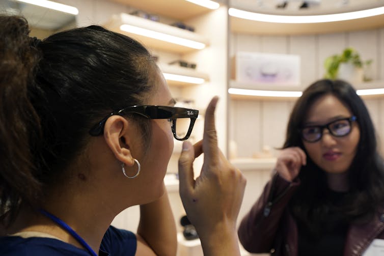 Two women with glasses on face each other in a store. One of the women is pointing to the outside of the glasses she is wearing