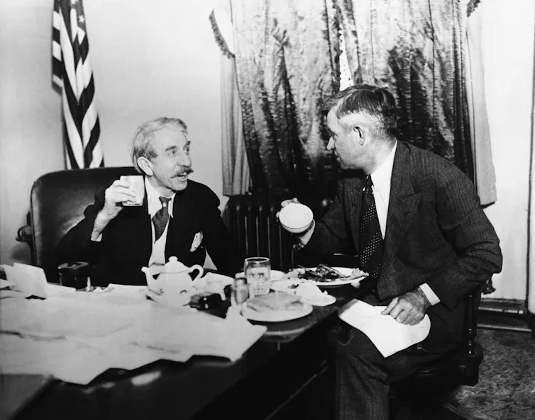 Two men eating at a desk with a flag behind them.