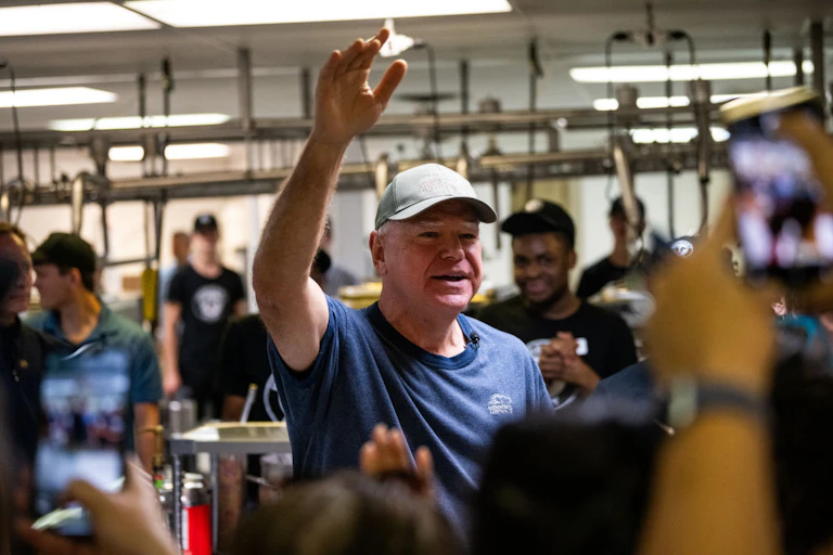 An older man wears a navy blue shirt and a grey hat and waves to people.