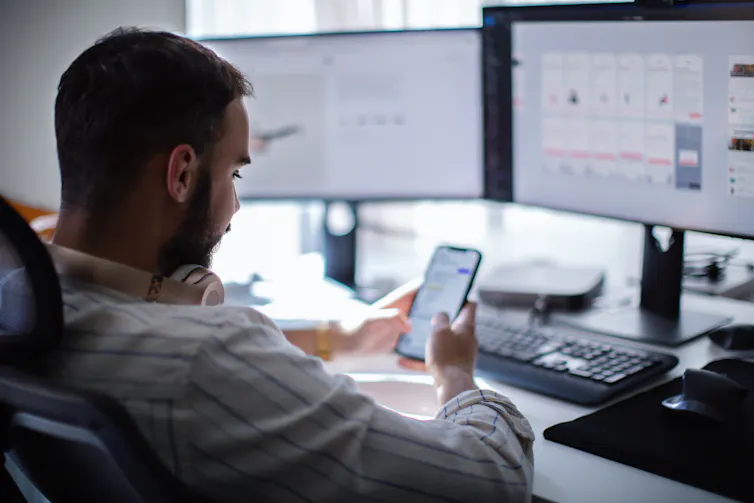 a man slouching in an office chair in front of a computer monitor looks at his phone