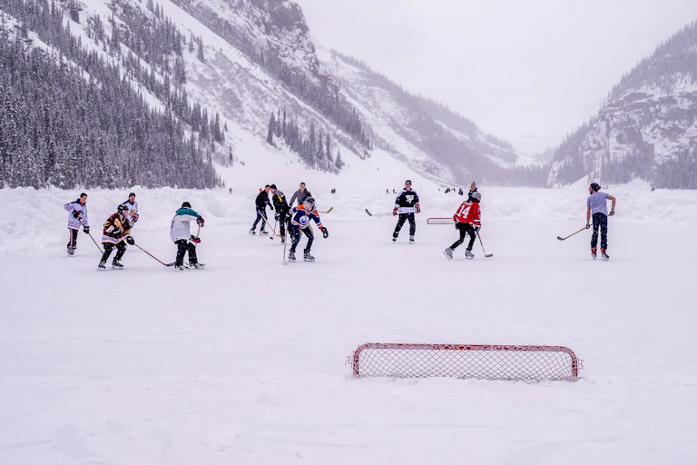 A group of people play hockey on a frozen ice pond with mountains in the background
