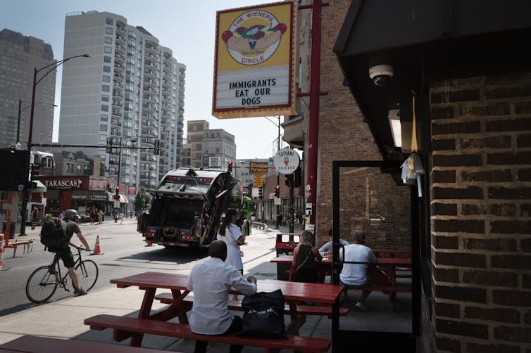 A person sits at a picnic table on a city street, below a sign that says 'The Wieners Circle' with a picture of a hot dog. It also says 'Immigrants eat our dogs.'