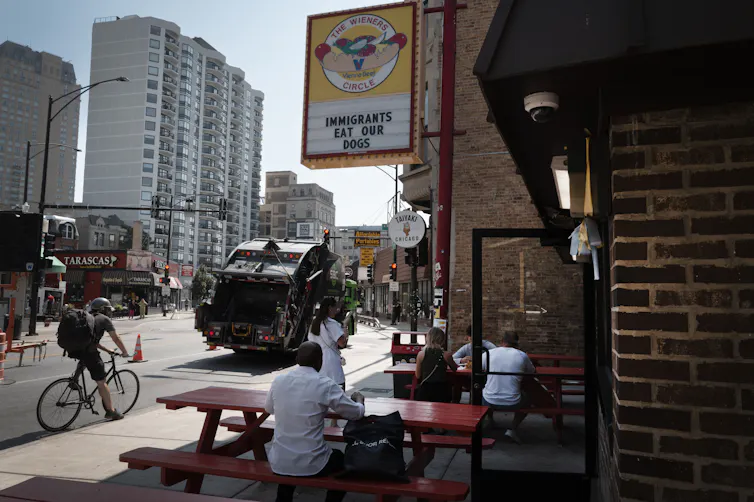 A person sits at a picnic table on a city street, below a sign that says 'The Wieners Circle' with a picture of a hot dog. It also says 'Immigrants eat our dogs.'