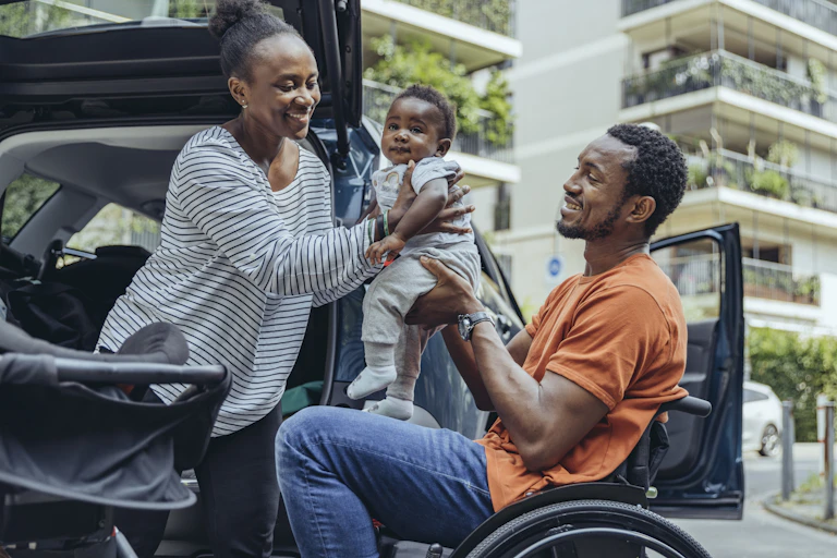 Mother giving son to father sitting in wheelchair by car