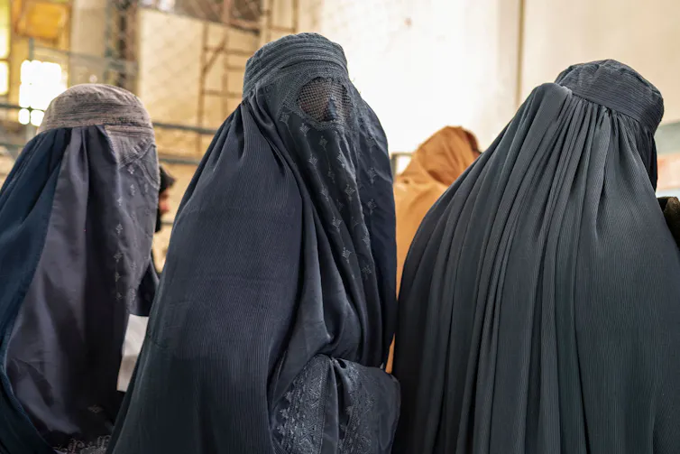 Mulheres usando hijab azul, que as cobrem da cabeça aos pés, ficam em uma fila.