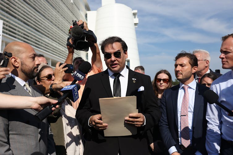 Santos stands in a scrum of reporters, holding a manila folder