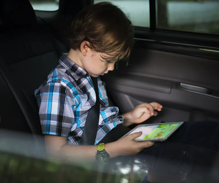 Little boy in a car wearing a plaid shirt and looking at a tablet in his lap