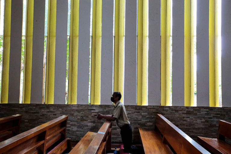 A man in a white shirt kneels at a pew in a darkened room with white slab walls, with greenery visible outside.