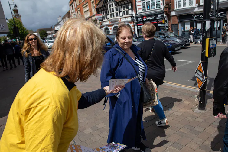 A woman in a yellow tshirt handing a leaflet to a passerby.